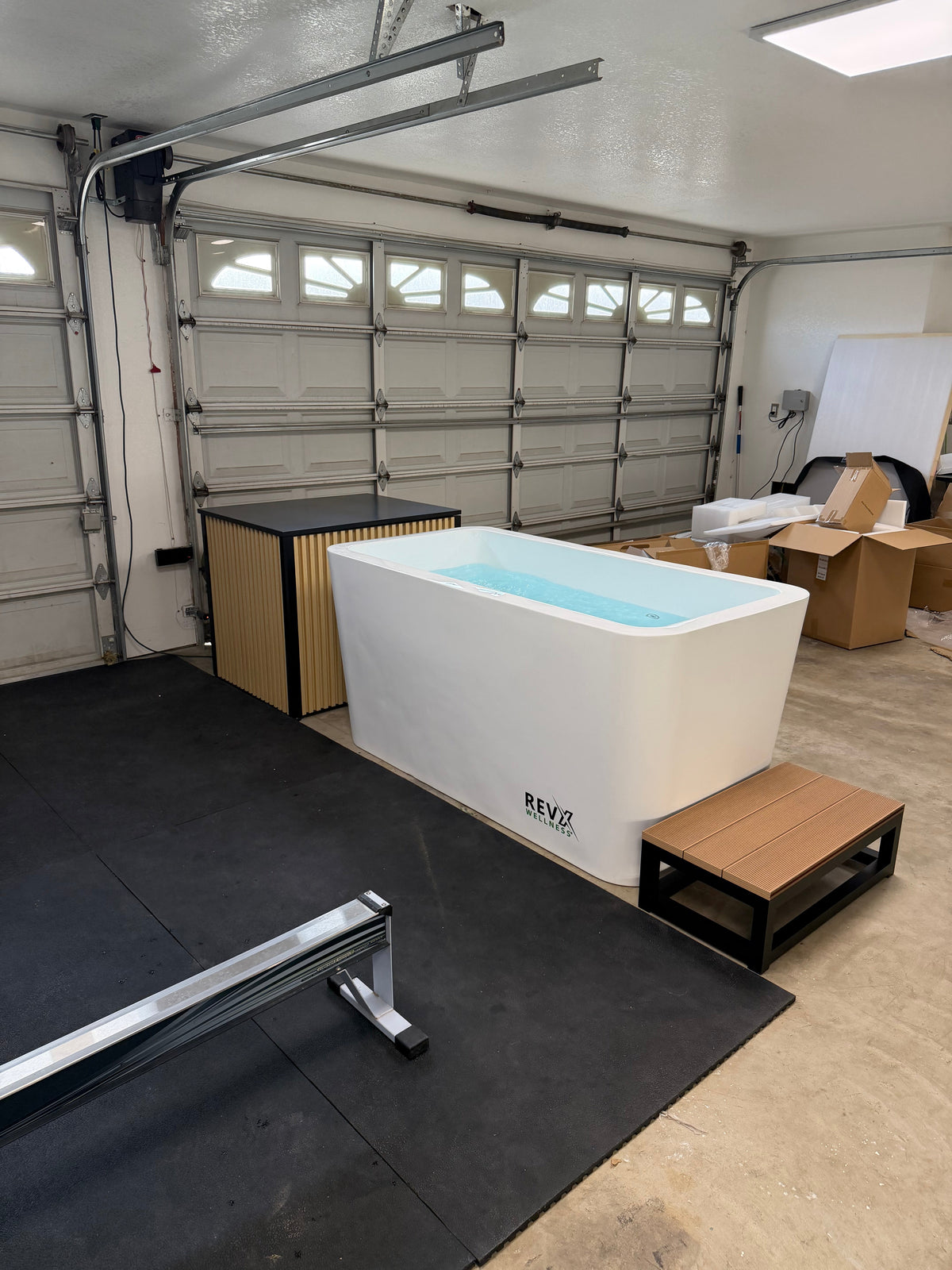 A freestanding rectangular bathtub with a light blue water fill, branded 'REVZ', shown in a garage setting with a wooden bench, black floor mat, and garage door background.
