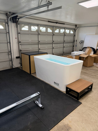 A freestanding rectangular bathtub with a light blue water fill, branded 'REVZ', shown in a garage setting with a wooden bench, black floor mat, and garage door background.