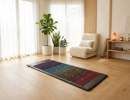 A rectangular massage mat with integrated LED light therapy panels lies on a light wood floor in a bright, modern room with a cream recliner, potted plants, and sheer curtains.